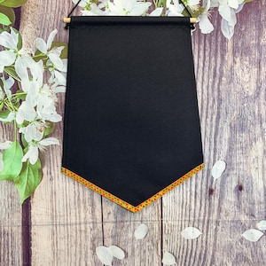 Puede incluir: Un tapiz de pared negro con un borde decorativo de triángulos rojos, amarillos y verdes. La pancarta está suspendida por un pasador de madera y una cuerda negra. El fondo es una superficie de madera con flores blancas.