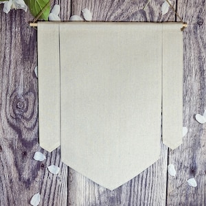 May include: A plain white canvas banner with a wooden dowel at the top and two strings hanging from the dowel. The banner is hanging on a wooden wall with white flower petals scattered around it.