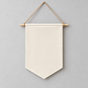 Op de afbeelding: Een blanco, gebroken wit canvas wandbanner met een puntige onderkant. De stof hangt aan een lichtgekleurde houten stok, die is bevestigd aan een dun, gedraaid bruin touw van jute om het aan een vlak oppervlak op te hangen.