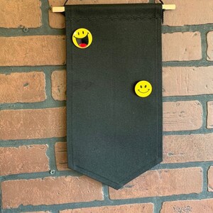 Puede incluir: Colgante de pared negro en forma de banderín con una clavija de madera y una cuerda. Dos imanes amarillos con caras sonrientes están adheridos a la tela. El fondo es una pared de ladrillos.