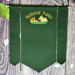 May include: A dark green banner featuring the embroidered text "National Parks" in yellow, and an image of a deer, trees, and mountains. The banner hangs from a wooden rod and has two vertical side panels.