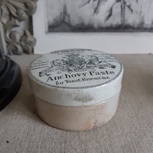 Puede incluir: Un tarro de cerámica blanca con una etiqueta en blanco y negro que dice "The Original Genuine Anchovy Paste for Toast, Biscuit &c." El tarro tiene un esmalte agrietado y está ligeramente astillado.