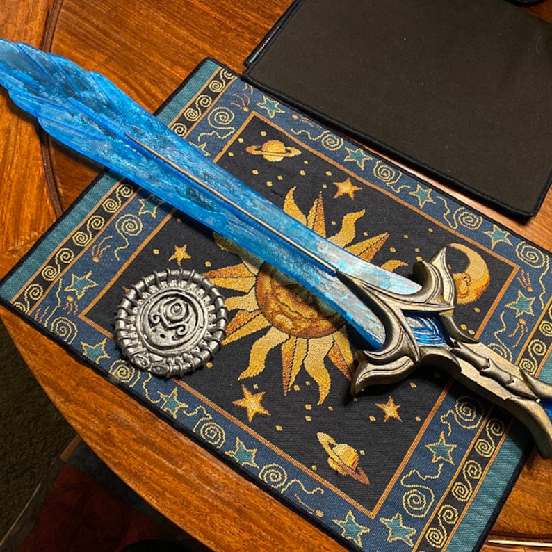May include: A blue and silver fantasy sword with a decorative handle, resting on a blue and gold tapestry with a sun and stars design.
