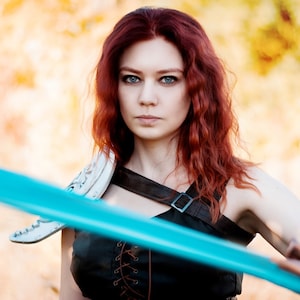 May include: A woman with long, curly red hair is holding a blue sword. She is wearing a black leather top with a white leather shoulder piece. The background is blurred and out of focus.
