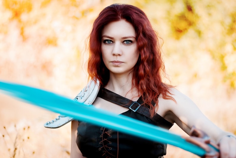 May include: A woman with long, curly red hair is holding a blue sword. She is wearing a black leather top with a white leather shoulder piece. The background is blurred and out of focus.
