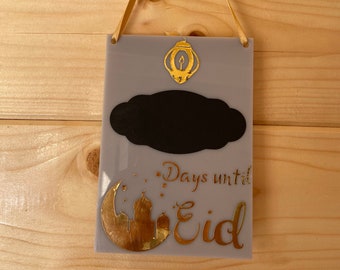 Puede incluir: Un letrero de madera gris con una luna creciente dorada, una mezquita y las palabras "Days until Eid" en letras doradas. Hay una pizarra negra en forma de nube con una linterna dorada encima.