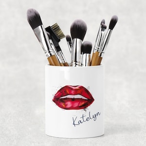 May include: White cylindrical makeup brush holder featuring a red lips design and the name "Katelyn". The holder contains makeup brushes with silver handles and black and brown bristles. The design is a beauty theme.