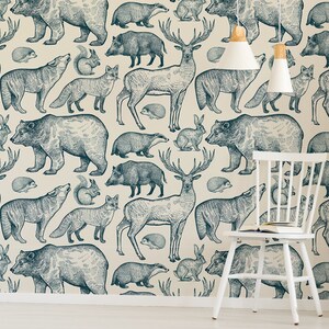 May include: A white wall with a blue and white patterned wallpaper featuring various woodland animals, including bears, deer, wolves, and foxes. The wallpaper is in a nursery or children's room with white furniture, including a white chair and two white bookshelves with wooden shelves. The bookshelves are filled with books, toys, and plants.