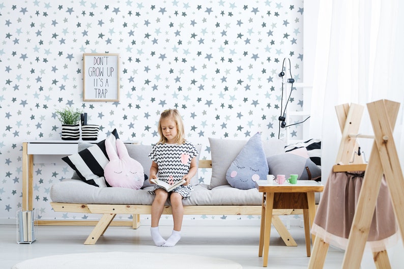 May include: A child sits on a gray couch with a gray and white star patterned wallpaper. The couch has a pink bunny pillow, a gray pillow with a raindrop design, and a black and white striped pillow. The child is reading a book. A framed sign on the wall reads "Don't grow up, it's a trap."
