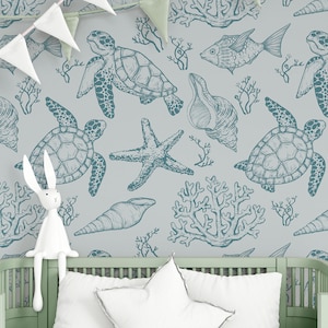 Op de afbeelding: Een lichtblauwe en witte babykamerbehang met een herhalend patroon van zeeschildpadden, zeesterren, schelpen en koraal. De behang is te zien op een muur achter een wit wiegje met een wit knuffelkonijn dat op de rand zit.