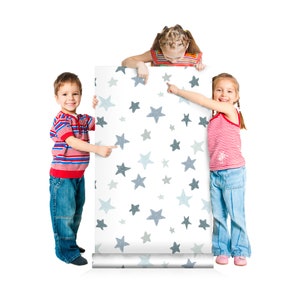 May include: Three children stand in front of a roll of wallpaper with a white background and a pattern of blue stars. The children are pointing at the wallpaper.