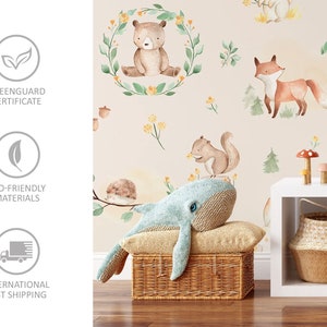 May include: A light beige wallpaper with a woodland animal theme. The wallpaper features a bear, fox, squirrel, and hedgehog. The wallpaper is shown in a nursery setting with a blue whale plush toy, a wicker basket, and a white shelf.