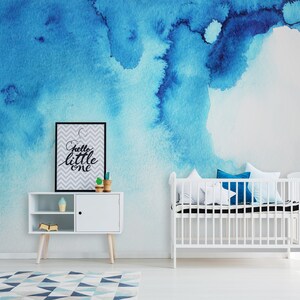 Puede incluir: Una cuna blanca con un colch&oacute;n blanco y almohadas azules en una habitaci&oacute;n de beb&eacute; con un mural de acuarela azul en la pared y un armario blanco con una impresi&oacute;n enmarcada en negro que dice "hello little one".