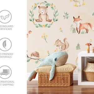 May include: A light beige wallpaper with a woodland animal theme. The wallpaper features a bear, fox, squirrel, and hedgehog. The wallpaper is shown in a nursery setting with a blue whale plush toy, a wicker basket, and a white shelf.