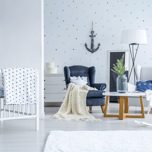 May include: A white crib with a blue and white patterned blanket, a blue and white patterned pillow, and a white wooden crate with a blue and white patterned pillow on top. There is a blue armchair with a white blanket, a white armchair with a blue pillow, a white rocking horse, a wooden coffee table, and a vase with green plants on the table. The wall is decorated with a blue anchor and white polka dots.