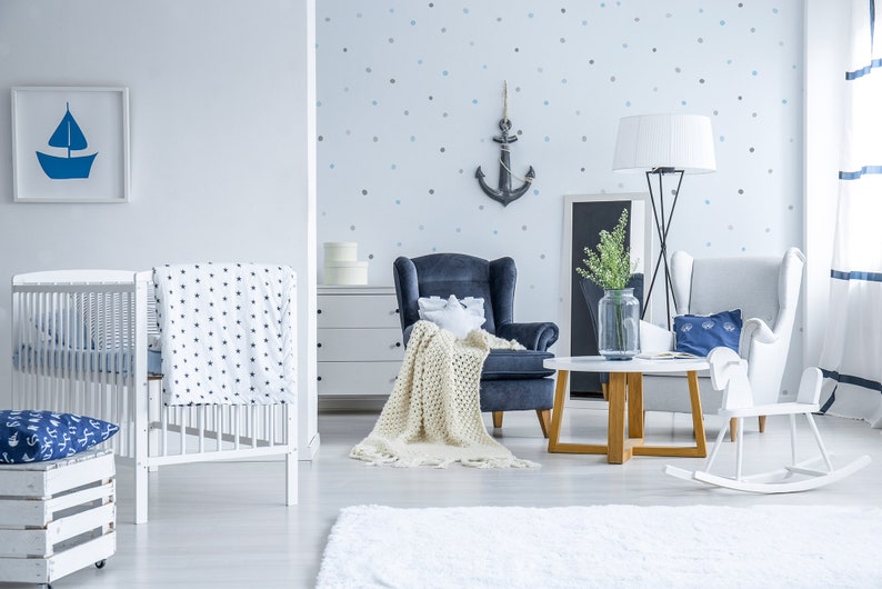 May include: A white crib with a blue and white patterned blanket, a blue and white patterned pillow, and a white wooden crate with a blue and white patterned pillow on top. There is a blue armchair with a white blanket, a white armchair with a blue pillow, a white rocking horse, a wooden coffee table, and a vase with green plants on the table. The wall is decorated with a blue anchor and white polka dots.