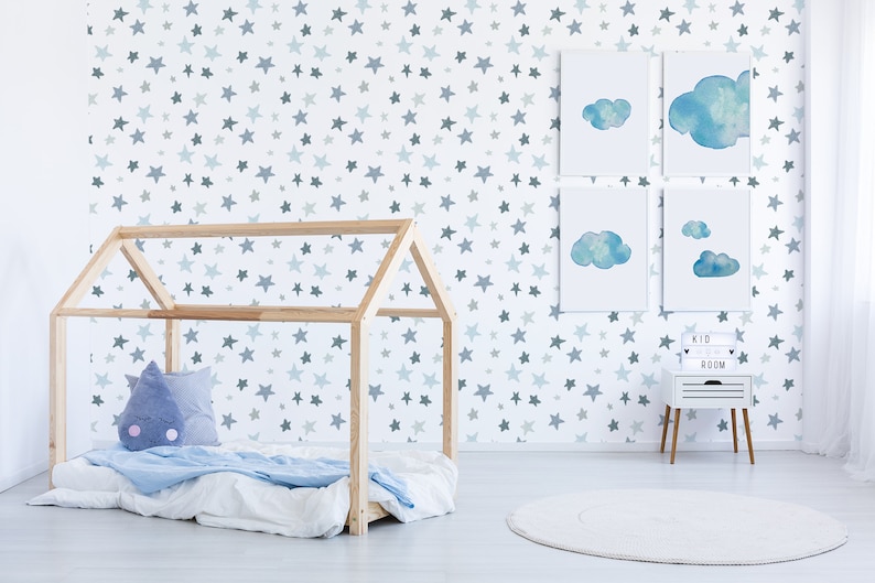 May include: A white wall with a blue and gray star pattern wallpaper. There is a wooden house-shaped bed frame with a blue and white blanket and a blue pillow with a cloud design. There are four framed watercolor paintings of clouds on the wall. A white nightstand with a sign that says "Kid Room" is next to the bed. There is a white round rug on the floor.