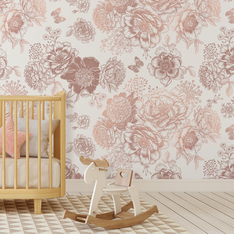 May include: A white nursery wall with a rose gold floral wallpaper featuring a repeating pattern of large and small flowers. A wooden cot with a pink pillow and a white rocking horse are in the foreground.