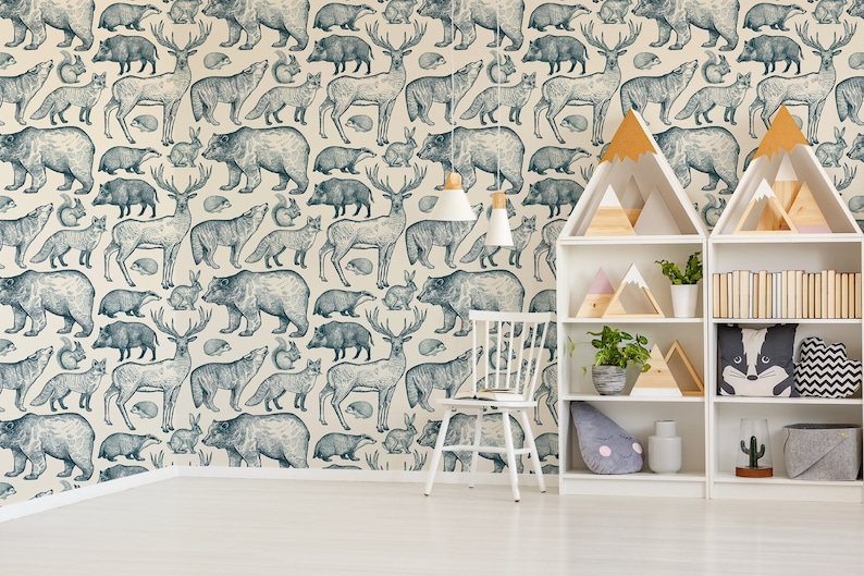 May include: A white wall with a blue and white patterned wallpaper featuring various woodland animals, including bears, deer, wolves, and foxes. The wallpaper is in a nursery or children's room with white furniture, including a white chair and two white bookshelves with wooden shelves. The bookshelves are filled with books, toys, and plants.