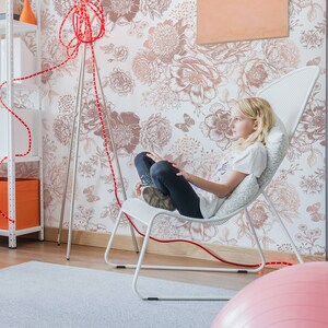 May include: A young person sits in a white mesh chair with a pink exercise ball in the foreground. The chair is in front of a wall with a floral wallpaper pattern. A red cord runs from the floor to the chair.