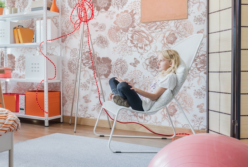 May include: A young person sits in a white mesh chair with a pink exercise ball in the foreground. The chair is in front of a wall with a floral wallpaper pattern. A red cord runs from the floor to the chair.