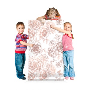 May include: Three children hold up a white and rose gold floral wallpaper roll. The wallpaper features a repeating pattern of delicate flowers and leaves.