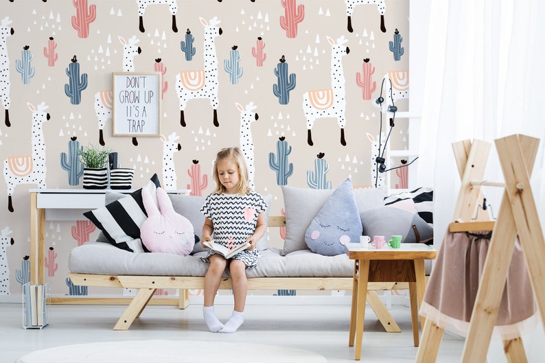 Puede incluir: Una habitaci&oacute;n infantil con papel pintado beige con motivos de llamas y cactus. Un letrero enmarcado dice "DON'T GROW UP IT'S A TRAP". Una ni&ntilde;a est&aacute; sentada en un sof&aacute; gris con cojines, leyendo. Tambi&eacute;n se ve una mesita auxiliar y un escritorio de madera.