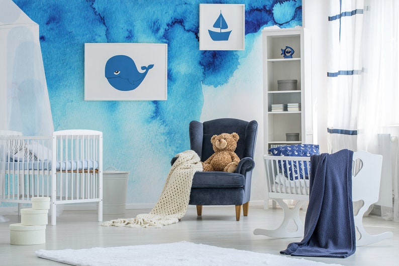 Puede incluir: Una habitaci&oacute;n de beb&eacute; con una pared con mural de acuarela azul que presenta una ballena y un velero. La habitaci&oacute;n tiene una cuna blanca, un mois&eacute;s mecedor, un sill&oacute;n azul con un oso de peluche y una estanter&iacute;a blanca con detalles azules.