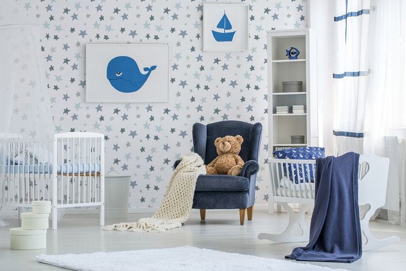 May include: A white crib with a blue and white patterned sheet, a white rocking bassinet with a blue and white patterned sheet, a blue armchair with a teddy bear sitting on it, and a white bookshelf with blue and white decorations. The room has a white wall with a blue star pattern.