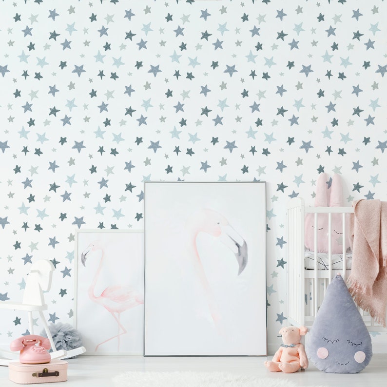 May include: A white wall with a blue watercolor star pattern. Two framed pictures of pink flamingos are hanging on the wall. A white crib with a pink bunny and a gray teardrop-shaped pillow is in the corner of the room.