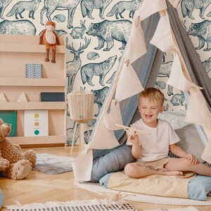 May include: A child sits inside a teepee tent in a room with a wallpaper featuring a repeating pattern of blue and white illustrations of various forest animals, including bears, deer, foxes, and squirrels. The room also features a wooden bookshelf with a stuffed animal on top, a blue rug, and a wooden floor.