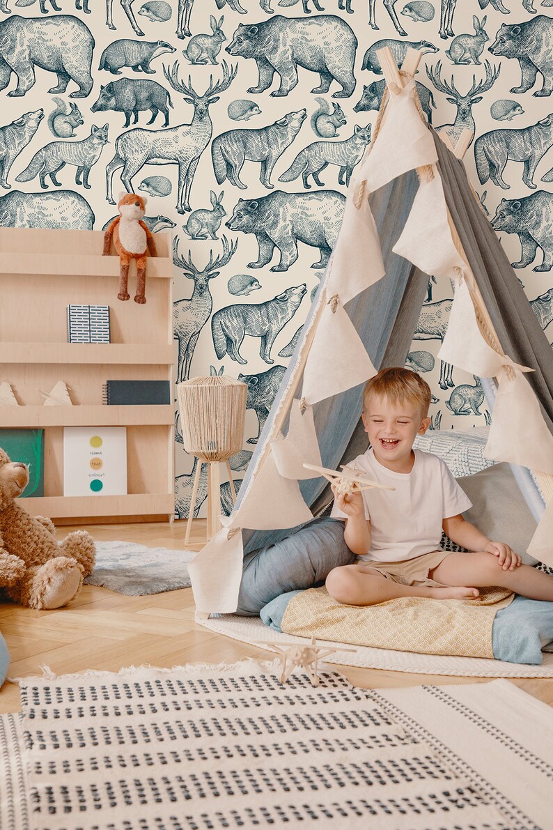 May include: A child sits inside a teepee tent in a room with a wallpaper featuring a repeating pattern of blue and white illustrations of various forest animals, including bears, deer, foxes, and squirrels. The room also features a wooden bookshelf with a stuffed animal on top, a blue rug, and a wooden floor.