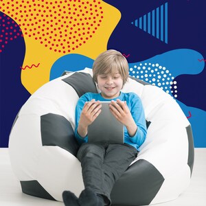 May include: A young person sits in a beanbag chair shaped like a soccer ball, using a tablet computer. The background is a colorful wall with geometric shapes and patterns.