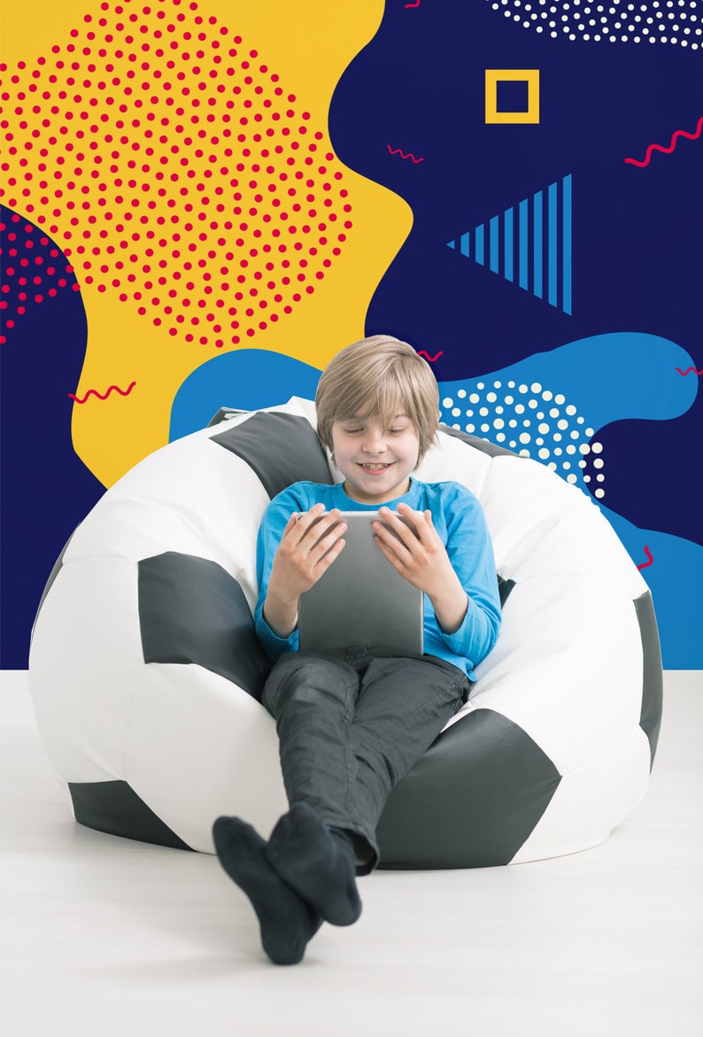 May include: A young person sits in a beanbag chair shaped like a soccer ball, using a tablet computer. The background is a colorful wall with geometric shapes and patterns.