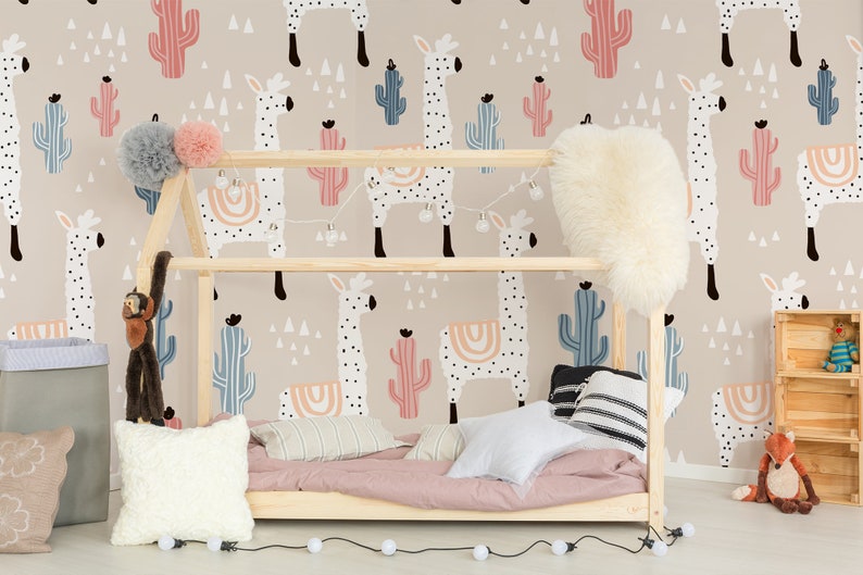 Puede incluir: Dormitorio infantil con papel pintado beige con motivos de llamas y cactus. Una estructura de cama de madera est&aacute; decorada con almohadas, una manta rosa y luces de cadena. Se ven un mono y un zorro de peluche.