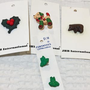 May include: A collection of decorative pins on white cards. The pins include a green holly with red berries, a brown bear, a tan dog with red and green accents, and two green frog pins. The cards have the text "JHB International" and "FUNTASTICS by Streamline".