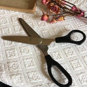 May include: A pair of black handled pinking shears with a silver blade. The shears are open and lying on a white fabric surface.