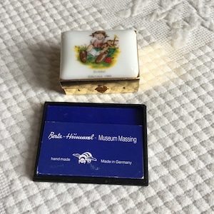 Puede incluir: Pequeña caja rectangular con marco de metal dorado y tapa de porcelana blanca. La tapa presenta una colorida ilustración de un niño con un oso de peluche. El texto "Hummel" y "Cöschmid 1984" están impresos en la tapa. Una tarjeta azul con texto está en la caja abierta.