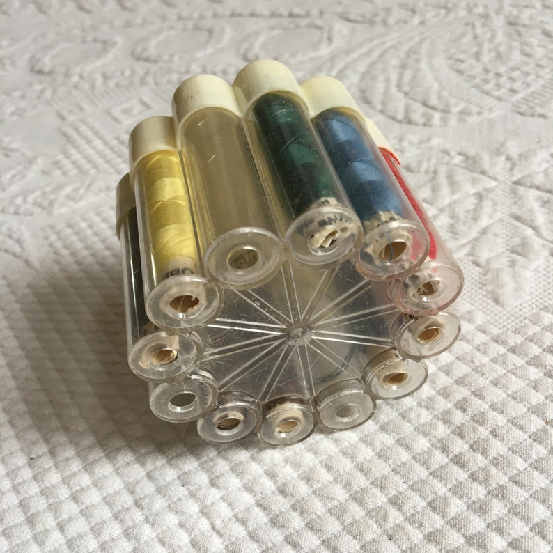 May include: A clear plastic container with 12 spools of thread. The spools are arranged in a circle and are visible through the clear plastic. The spools are in various colors, including yellow, green, blue, and red.