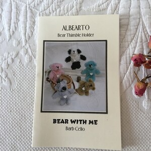 May include: A sewing pattern for a bear-shaped thimble holder. The pattern is titled "ALBERTO Bear Thimble Holder" and "BEAR WITH ME Barb Celio". The pattern includes a photo of five different bears, each with a different color and design.