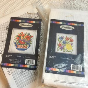 May include: Two Herrschners cross-stitch trivet kits. One features a floral design in a blue teapot, the other a bouquet of spring flowers. The kits include the fabric, pattern, and instructions.