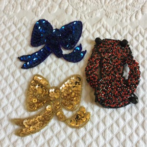 May include: Three decorative fabric appliques featuring bows made of sequins. One bow is blue, one is gold, and one is red and black.