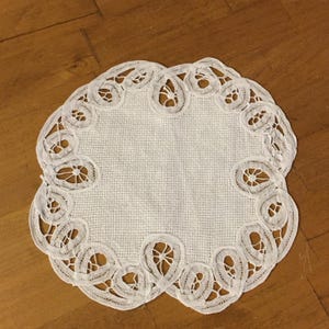 May include: A white, round doily with a delicate, openwork lace border. The center is a solid, textured fabric. The lace design features a repeating pattern of teardrop shapes with intricate cutouts, creating an elegant and vintage aesthetic.