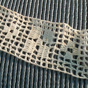 May include: Close-up of a cream-colored, open-weave crochet lace trim. The lace has a repeating geometric pattern and is set against a blue and white striped fabric background. The trim appears to be vintage or handmade.