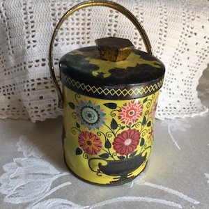 May include: A yellow metal tin with a black lid and a gold handle. The tin is decorated with colorful flowers and a black and gold design.