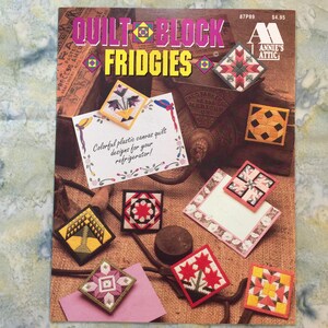 May include: A craft pattern booklet titled Quilt Block Fridgies by Annie's Attic featuring various square plastic canvas quilt designs. The cover displays multiple colorful stitched squares with geometric and floral patterns on a brown background.