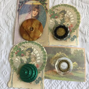 May include: A collection of vintage buttons in various colors and designs, including a large brown button, a black and gold button, a green button, and a clear button with a gold center. The buttons are displayed on antique postcards.