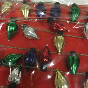 May include: A close-up of a string of Christmas lights. The lights are teardrop-shaped and come in various colours, including green, red, silver, gold, and blue. The lights are strung on a green wire against a red background.