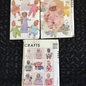 May include: Three vintage sewing pattern envelopes featuring baby doll clothing designs. The patterns showcase various outfits in pastel colors, including dresses, jumpsuits, and hats. The McCall's Crafts pattern includes multiple doll images.