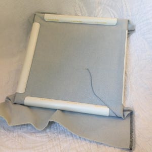 May include: A light blue fabric stretched over a white plastic frame. The fabric is held in place by white plastic clips. A needle and thread are visible in the center of the fabric.